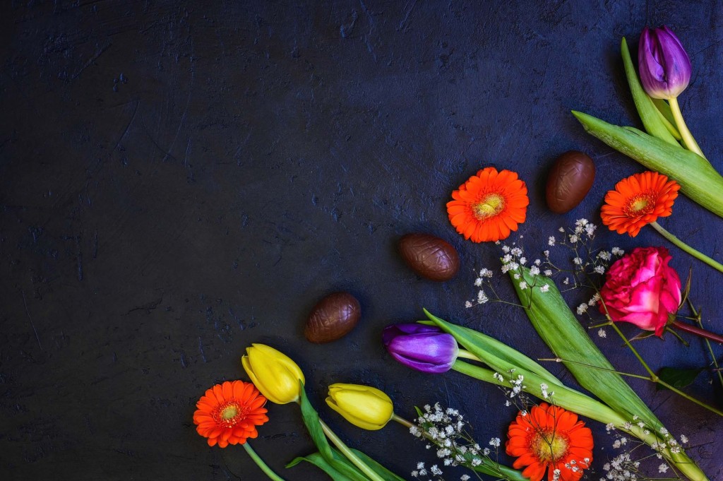 Flat lay composition with easter chocolate eggs and spring colorful flowers on dark blue rustic background. Happy Easter greeting card.