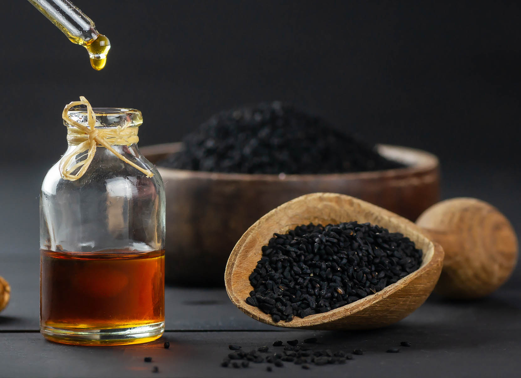 Glass bottle of black cumin seed essential oil , Nigella Sativa in scoop on black wooden background, oil dripping