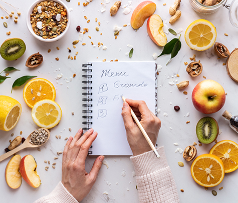 Nutritionist female hand write in notebook on healthy food background, women diet nutrition recipe menu, fresh summer fruit granola seeds on white table organic super food health care detox, top view
