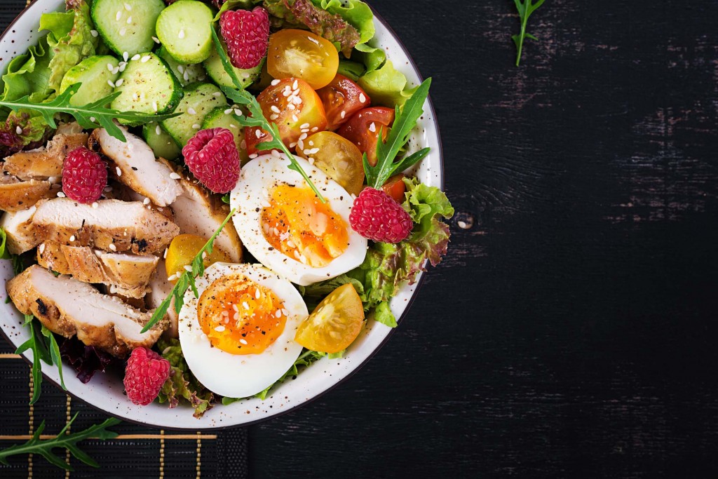 Grilled chicken meat and fresh vegetable salad of tomato, cucumber, egg, lettuce and raspberry. Ketogenic diet. Buddha bowl dish on dark background. Top view, flat lay
