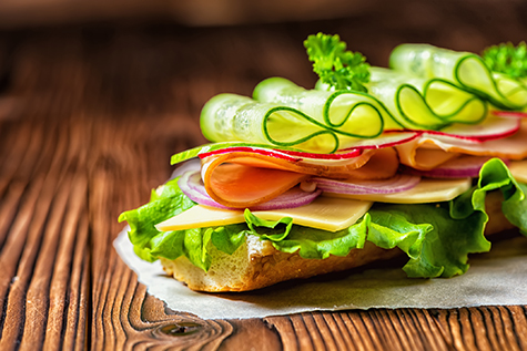 close up of sandwich with ham, cheese, bacon, radish, lettuce, cucumbers and onions on paper, wooden background