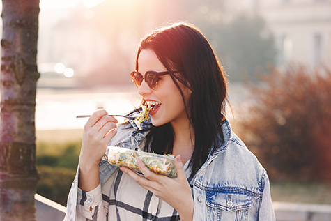 Young brunette woman in sunglasses eating fresh salad outdoor in sunset, looking away