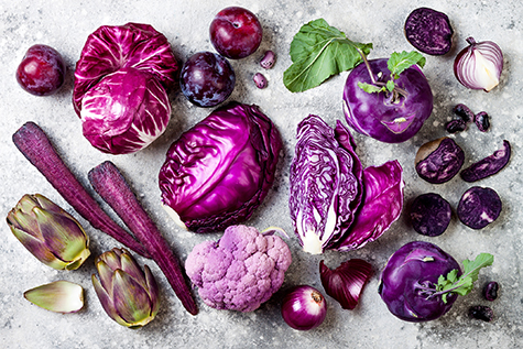 Raw purple vegetables over gray concrete background. Cabbage, radicchio salad, olives, kohlrabi, carrot, cauliflower, onions, artichoke, beans, potato, plums. Top view, flat lay.