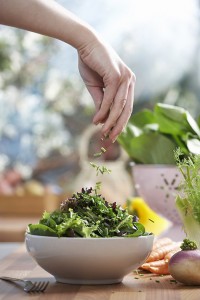 Woman pouring herbs into bowl of salad in kitchen, close-up of hand