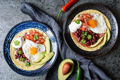 Huevos Rancheros Tostadas, Mexican breakfast consisting of toasted tortilla, chopped tomato and onion, jalapeno and beans, topped with fried egg, served with avocado slices and sour cream